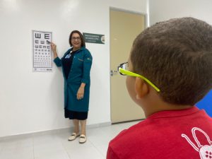 A imagem mostra uma criança que usa óculos sentada diante de uma parede que conta com elementos para realização de teste de aptidão visual.